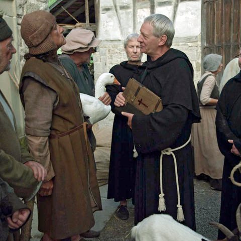 Auf einem Marktplatz steht eine Gruppe von Bauern drei Kleriker gegenüber. Die Kleriker tragen dunkle Kutten, einer von ihnen trägt eine Bibel. 