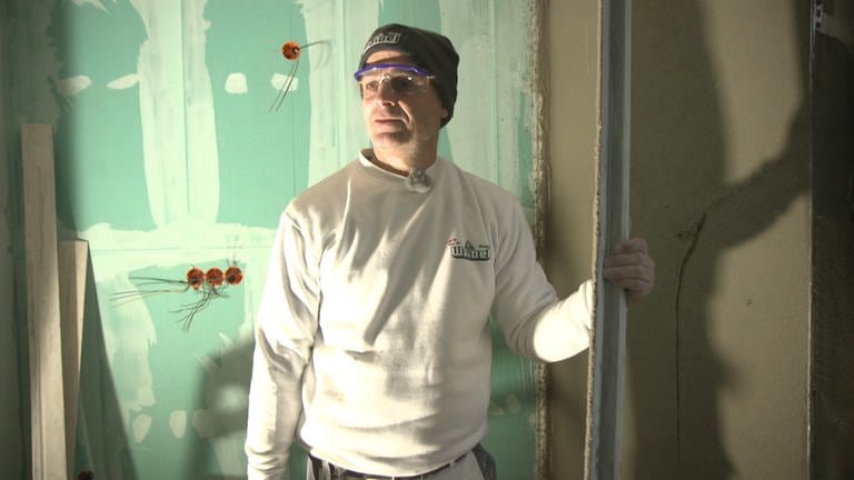 Stuckateur Manfred ist auf einer Baustelle. Er trägts seine Arbeitskleidung und eine Schutzbrille. Im Hintergrund stehen Kabel aus den Wänden. 