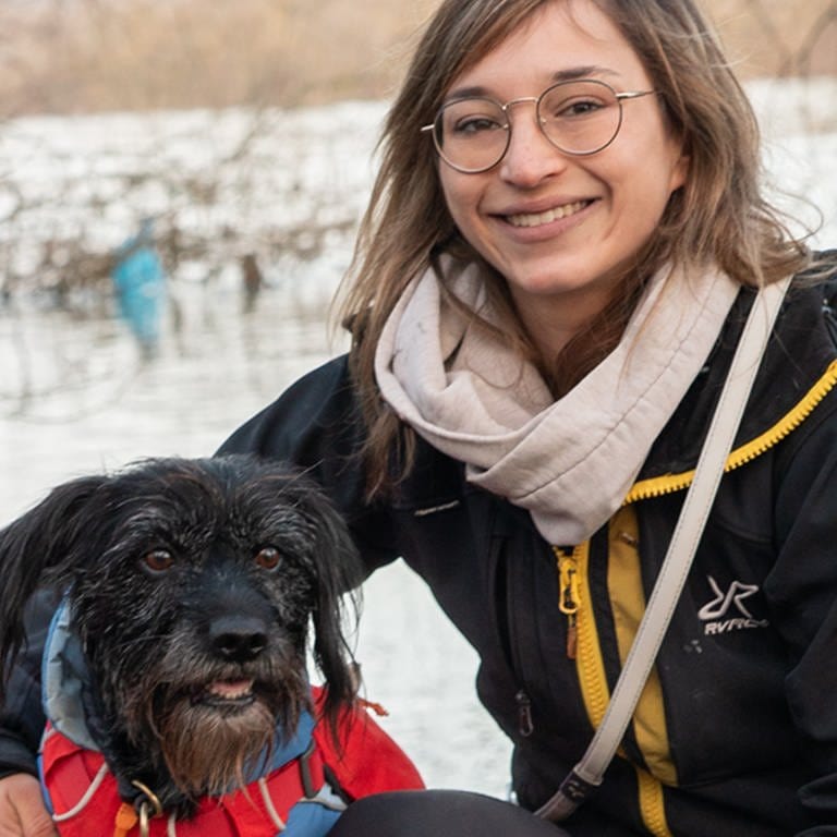 Hund und Frau mit Brille lächeln vor Fluss.
