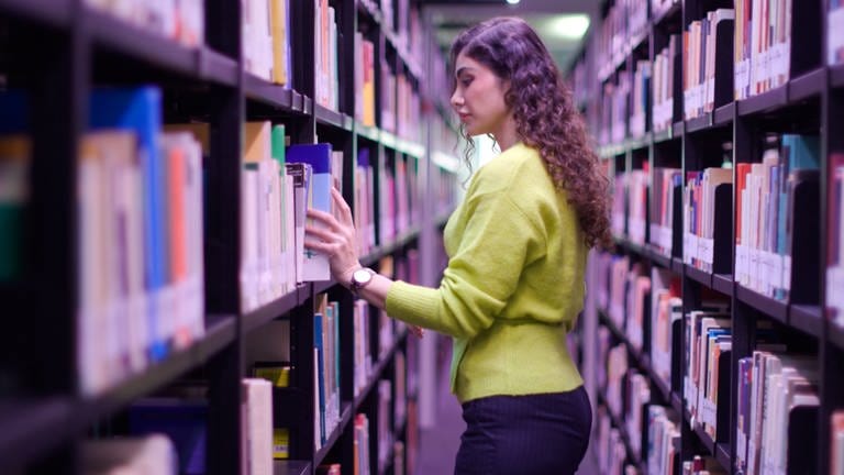  Eine Frau mit dunkelhaarigen Locken und einem gelben Pulli steht seitlich zur Kamera gedreht zwischen zwei Bücherregalen in einer Bibliothek und nimmt sich ein Buch heraus.