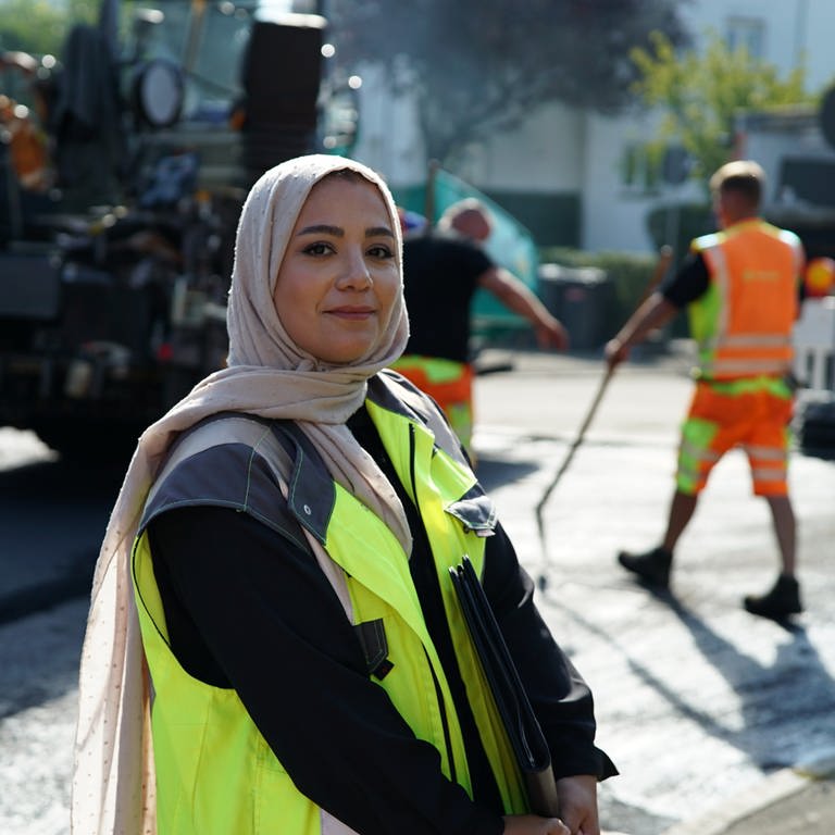 Bauingenieurin Hawra lächelt in die Kamera und steht auf einer Baustelle. Im Hintergrund sind Straßenbauarbeiter zu sehen.