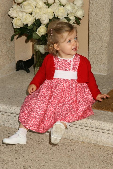 Die spanische Prinzessin Leonor sitzt als kleines Mädchen von einem Jahr in einem rotgeblümten Kleidchen und mit blonden Locken auf einer Treppenstufe vor einer Vase mit weißen Rosen und schaut zur Seite weg.