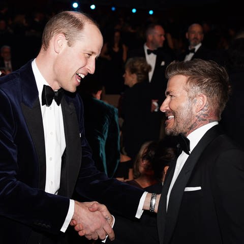 Prinz William trägt einen blauen Samtanzug mit weißem Hemd und einer passenden Schleife und schüttelt die Hand von dem britischen Fußballstar David Beckham, der einen schwarzen Anzug, ein weißes Hemd und eine Fliege trägt. Die beiden unterhalten sich vor der Verleihung der Bafta Film Awards 2024 in der Royal Festival Hall.