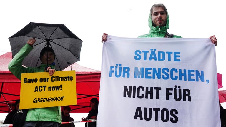 Zwei Demonstranten von Fridays For Future tragen Regenkleidung und halten Schilder und einen Regenschirm in der Hand.