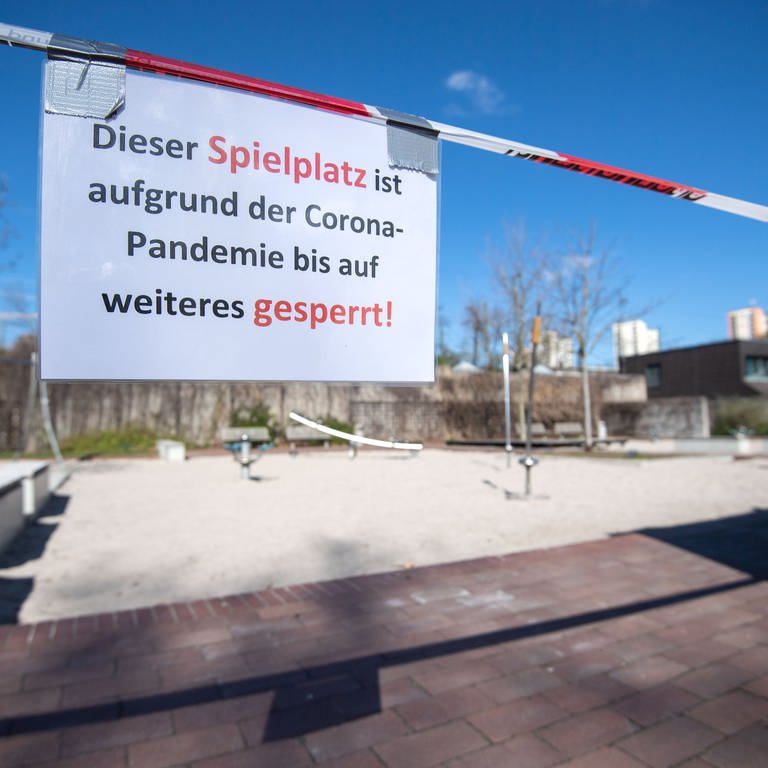 Ein Schild mit der Aufschrift "Dieser Spielplatz ist aufgrund der Corona-Pandemie bis auf weiteres gesperrt" hängt am Rand eines Spielplatzes an einem Absperrband.