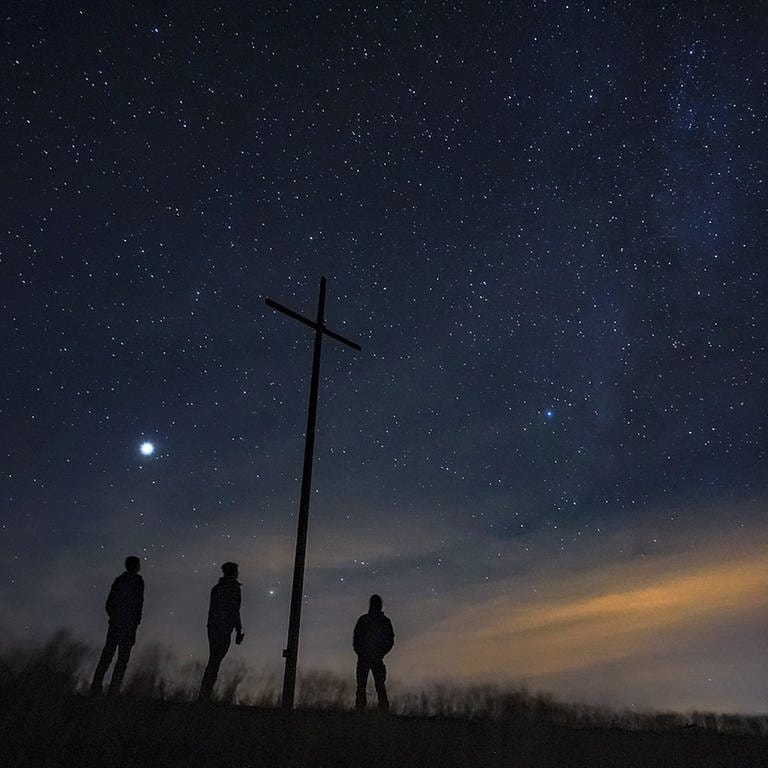 Drei Menschen blicken von einer Bergkuppe in einen Nachthimmel voller Sterne