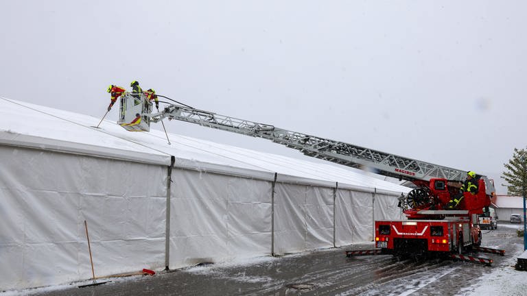 Feuerwehrleute stehen im Korb einer Drehleiter über dem Dach des Festzeltes in Riedlingen-Zell (Kreis Biberach).  Sie kehren Schnee vom Zelt. 