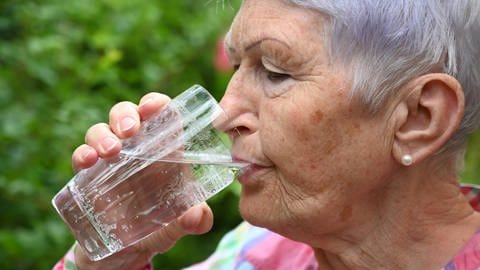 Ältere Frau trinkt Wasser aus einem Glas: Zellen brauchen Wasser, um richtig zu funktionieren. Gerade ältere Menschen trinken aber oft nicht ausreichend. Es ist aber vor einer OP besonders wichtig, dass Patienten gut mir Wasser versorgt sind – sogar bis unmittelbar vor der Operation. Das trägt dazu bei, ein postoperatives Delir zu vermeiden. Bisherige Standards sollten sich in den Kliniken ändern.